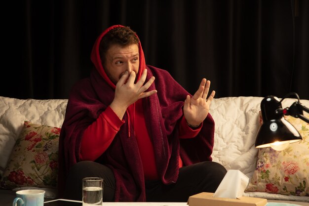 Man wrapped in a plaid looks sick, sneezing and coughing sitting at home indoors