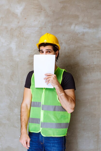 Man in yellow helmet and gear holding project reports and hiding his face behind it