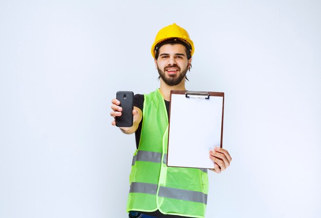 Man in yellow helmet holding a project folder and a smartphone.