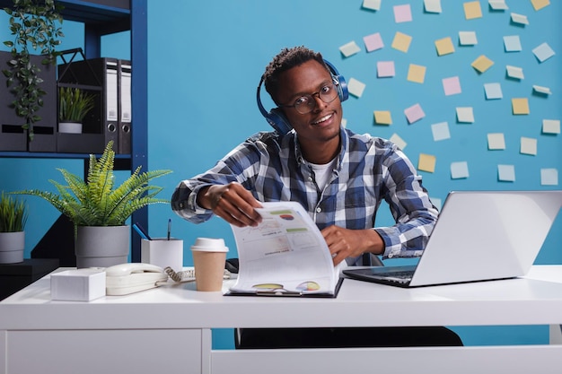 Management department team leader sitting at desk with financial charts while listening music. Young adult office worker smiling at camera while analyzing startup project promoting campaign.