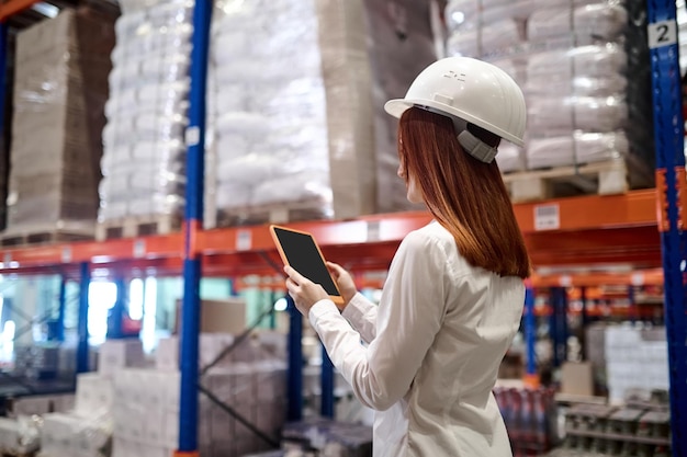 Free photo management, warehouse. profile of woman with long red hair in protective helmet looking at tablet screen walking along warehouse aisle