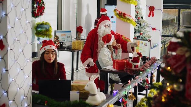 Free photo manager dressed as santa in xmas ornate office supervising tasks after offering presents to staff members team leader in festive workspace wearing christmas costume examining work done by employees