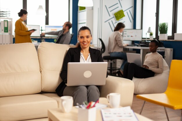 Manager lady writing on laptop looking at camera smiling while diverse colleagues working in background