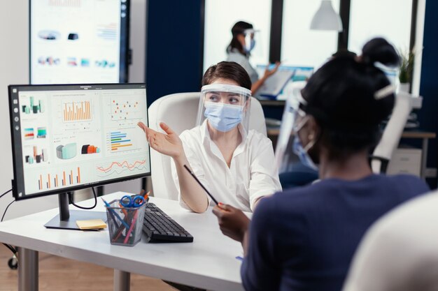 Manager wearing face mask for covid19 explaining financial graph to african employee. Multiethnic team working in company with new normal respecting social distance because of global pandemic with cor