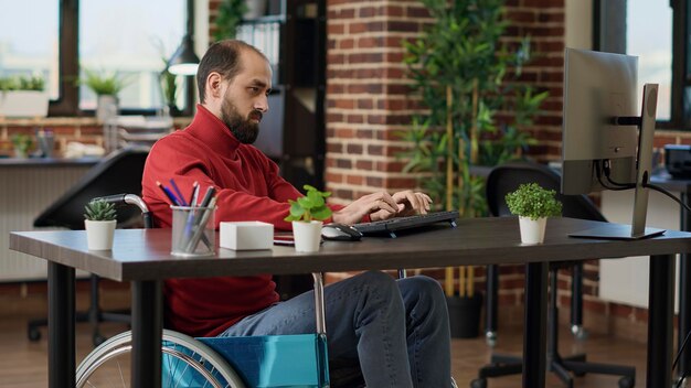 Менеджер с инвалидностью, работающий над развитием бизнеса с компьютером за столом, сидит в инвалидной коляске. Работник мужского пола планирует финансовую стратегию для создания исполнительной компании развития.