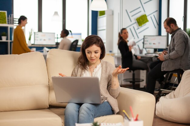 Manager woman sitting on couch holding laptop and talking on video call during virtual conference working in business modern office