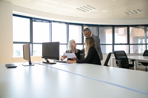 Managers discussing reports with boss, sitting at meeting table with monitor and holding documents. Business meeting or teamwork concept