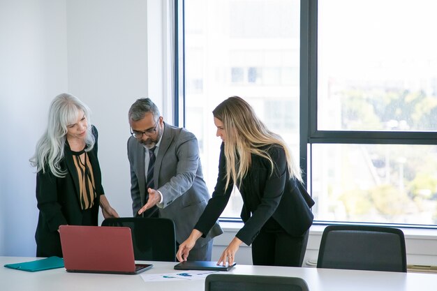 Managers showing project presentation to boss. Businessman pointing hand at laptop screen and talking to female colleagues. Wide shot. Business communication concept