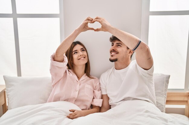 Mand and woman couple doing heart gesture sitting on bed at bedroom