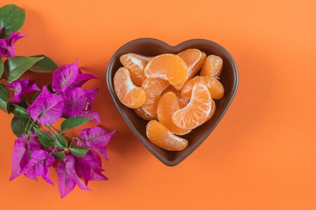 Mandarin in heart plate near the purple flower on orange surface