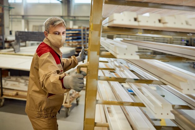 Manual worker with face mask moving rack with processed wood at carpentry workshop