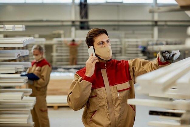 Manual worker with face mask talking on cell phone while working at carpentry workshop