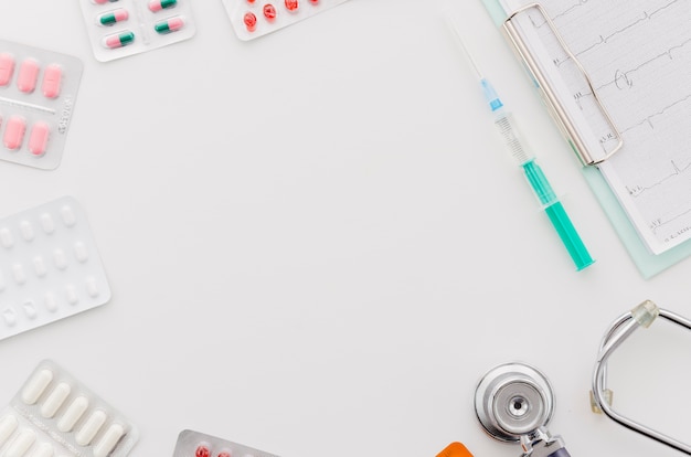 Many blister pills pack with syringe; stethoscope and ecg report on clipboard on white backdrop