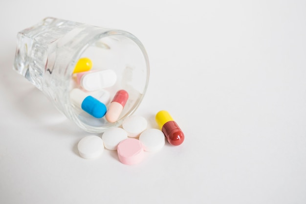 Many colorful pills spilling from tiny glass on white background