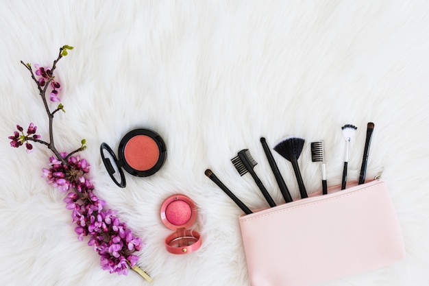 Many makeup brushes out of pink bag; flower twig and compact face powder on soft white fur