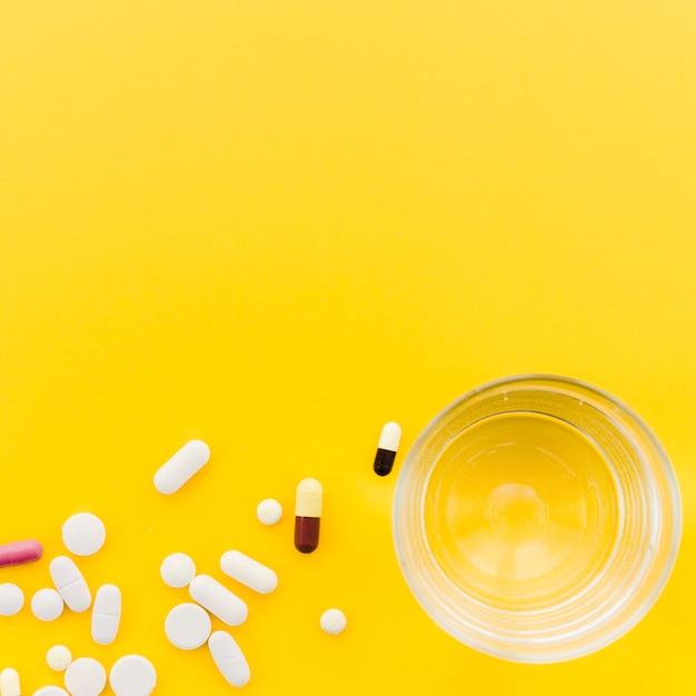 Many pill and capsules near the glass of water on yellow backdrop