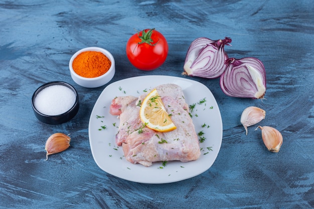 Бесплатное фото Маринованное куриное мясо и лимон на тарелке рядом с чесночным луком, помидорами и мисками для специй на синей поверхности