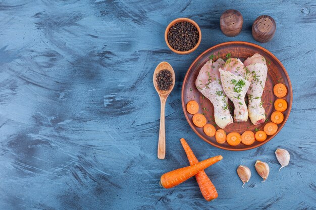 Marinated drumsticks and sliced carrots on a wooden plate next to spice, spoon and garlic, on the blue background. 