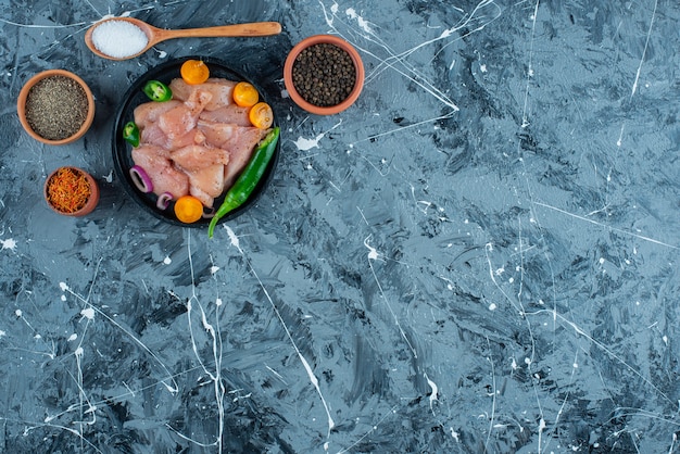 Carne e verdure marinate su un piatto accanto alle spezie in ciotole e cucchiaio sulla superficie blu