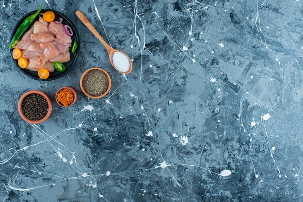 Marinated meat and vegetables on a plate next to spice in bowls and spoon on the blue surface