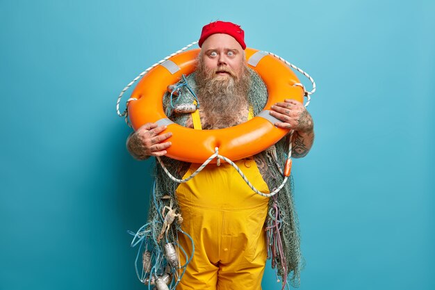 Marine profession. Stunned bearded seafarer stares has bugged eyes, poses with inflated swim ring, wears yellow overalls