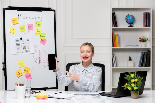 Маркетинговая умная милая бизнес-леди в полосатой рубашке в офисе держит мобильный телефон и улыбается