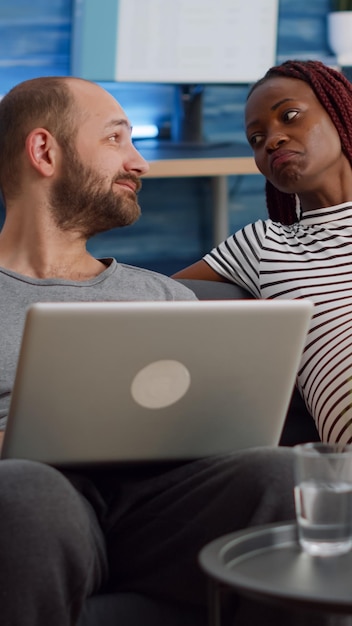 Married interracial couple talking about pregnancy while relaxing together in living room. multi ethnic people using laptop and watching television. black woman holding tv remote control