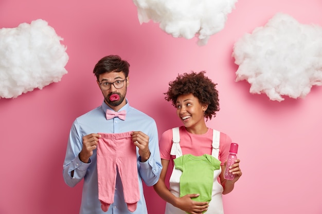 Free photo married mixed race couple expect baby, buy necessary items for newborn. cheerful pregnant woman holds bodysuit and feeding bottle for infant, looks gladfully at husband. happy expectations concept