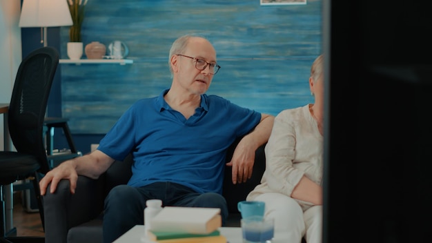 Married people watching movie on television in living room, sitting together and enjoying retirement at home. Senior couple looking at comedy on tv to do fun activity for entertainment.