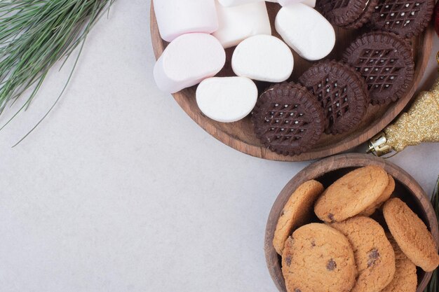 Marshmallows, cookies on wooden board on white table.