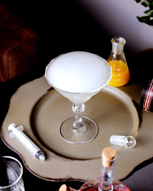A martini glass with smoked cocktail placed next to needle and conical flask with orange juice