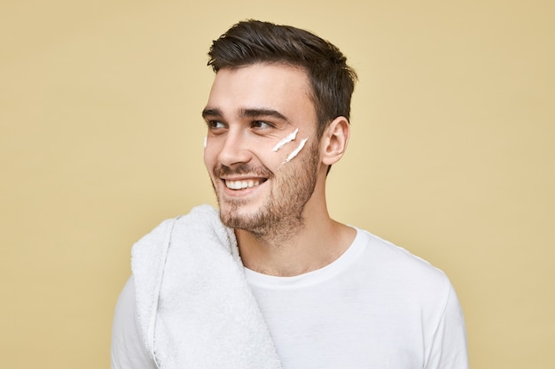 Masculine beauty concept. Happy attractive young Caucasian male with bristle posing in bathroom in the morning with towel on shoulder, looking away with smile, after shave cream on his face