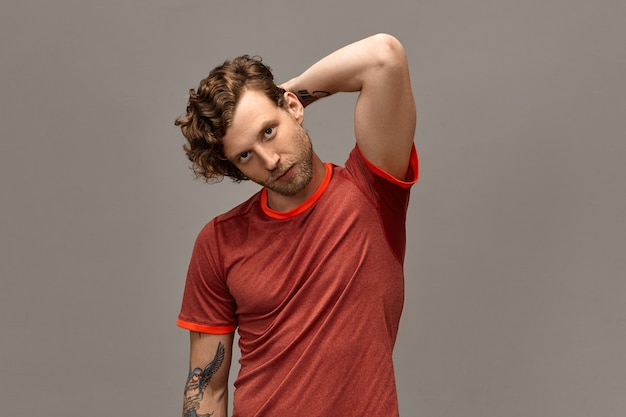 Masculine beauty, healthy active lifestyle and people concept. Portrait of confident good looking male runner with trimmed beard, curled hair and tattoo posing isolated, holding hand behind his head