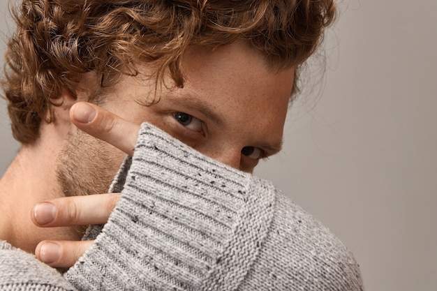 Masculine beauty, style and fashion concept. Close up of mysterious handsome young male with curly hair, beautiful magnetic eyes, his fingers stick out of gray knitted sleeve