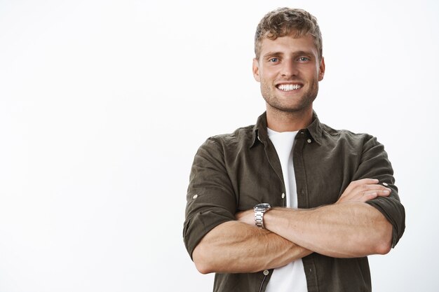 Masculine and strong good-looking man with white perfect smile and blue eyes cross hands against body in confident and assertive pose, satisfied and assured everything great over gray wall