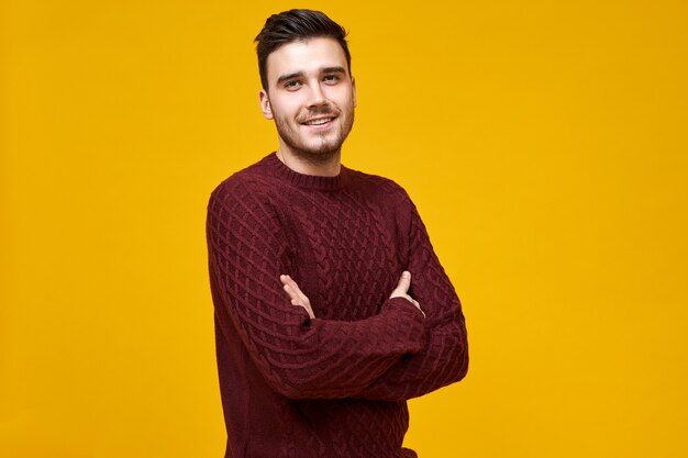Masculinity, confidence and body language. Isolated shot of handsome joyful young man in knitted maroon jumper feeling confident and proud of himself, keeping arms folded on her chest, smiling