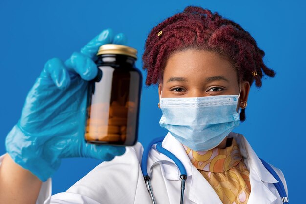 Masked african american woman doctor showing glass jar of medicine against blue background