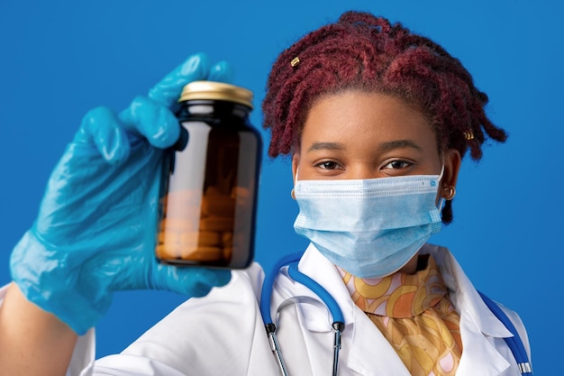 Masked african american woman doctor showing glass jar of medicine against blue background