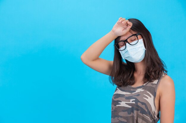 The masked girl shows her hand with her forehead closed on a blue wall.