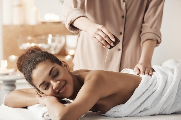 Masseur pouring oil doing massage for beautiful african woman in spa salon.