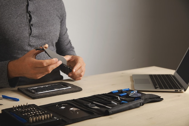 Free photo master holds new screen for replacement above disassembled smartphone in his laboratory, tool kit with instruments and laptop in front of him on white table, space for your text on right