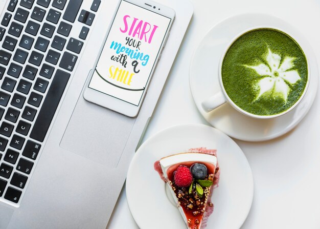 Matcha green tea latte cup; cheesecake and mobile phone with morning message on an open laptop on white desk