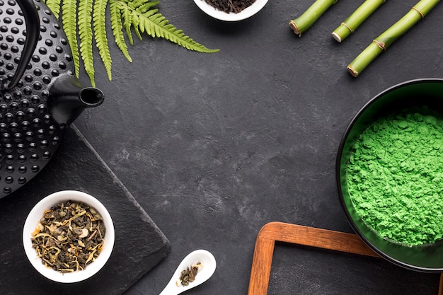 Matcha tea powder; dry herb; teapot; fern leaves and bamboo stick on slate stone backdrop