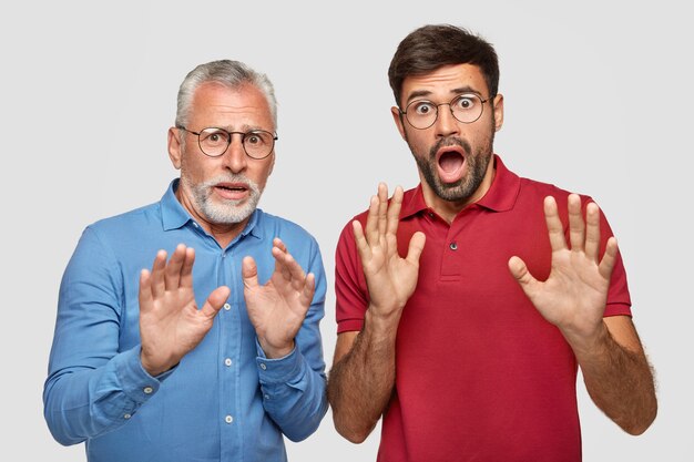 Mature and adult men have frightened expressions, see something scary in front, gesture with hands as try to defense, wears round glasses, stare with eyes popped out, isolated over white wall