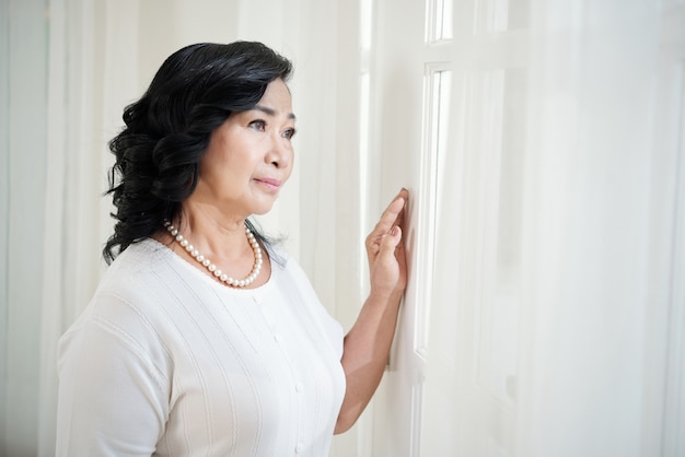 Зрелая азиатская дама готовя окно и смотря вне