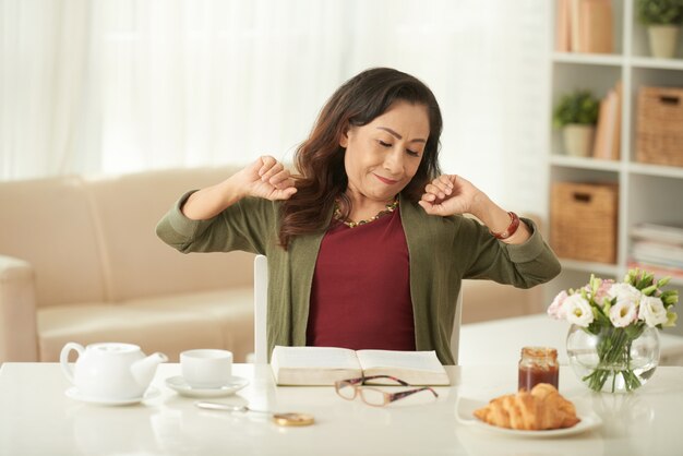 아침에 아침 식사 테이블에 앉아있는 동안 스트레칭 성숙한 아시아 여자