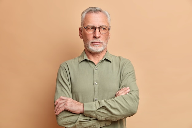 Mature bearded man executive keeps arms folded and stands thoughtful indoor thinks over future plans dressed in formal clothing isolated over beige wall needs to thinks about something