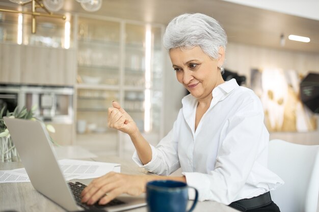 mature businesswoman using laptop for remote work, sitting at desk with coffee
