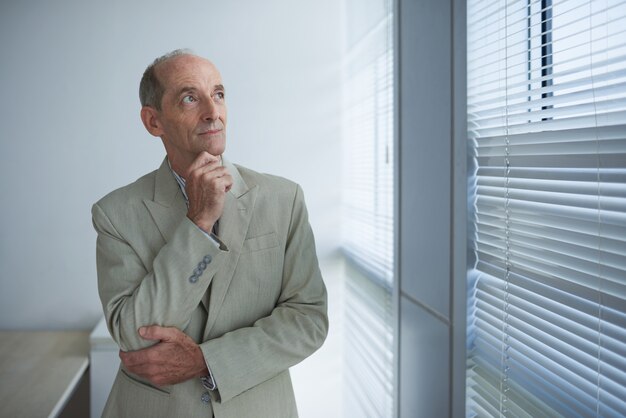 Mature Caucasian businessman in suit standing near window with closed blinds and looking away