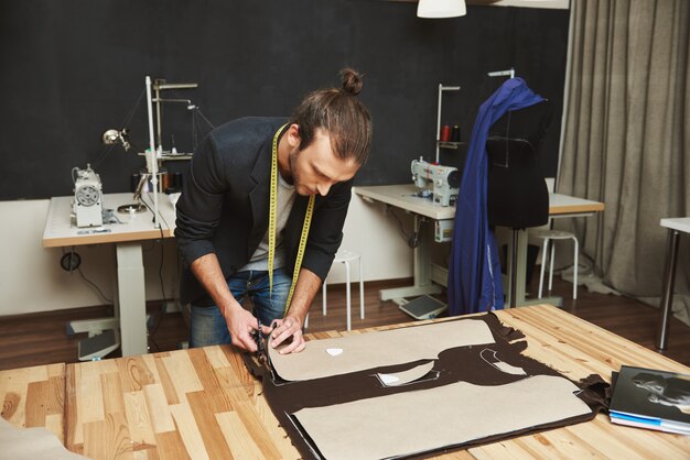Зрелый творческий красивый темноволосый кавказских мужчин модельер в черном костюме, вырезать одежду частей из ткани с ножницами, проводя вечер в студии.